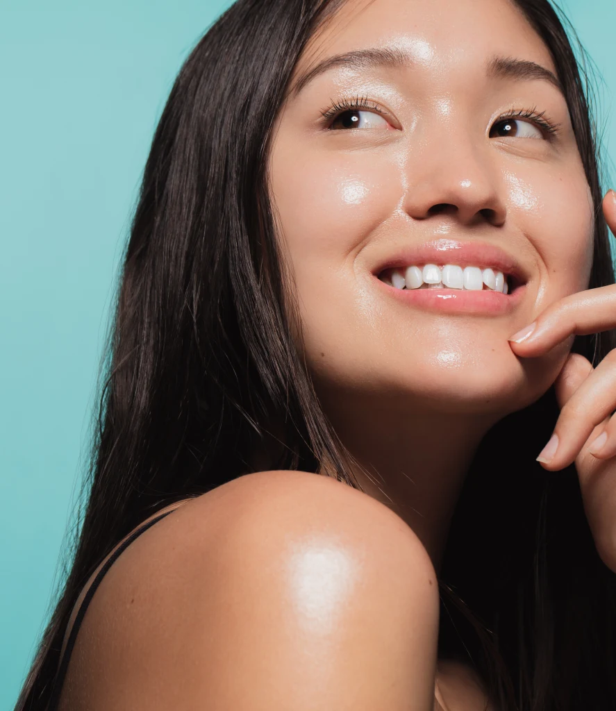 Smiling woman with radiant, glowing skin touches her chin, showcasing the results of PRP Microneedling Treatment Three in Centennial CO at Refine Med Spa. Her clear complexion and healthy glow emphasize the rejuvenating effects of the procedure.